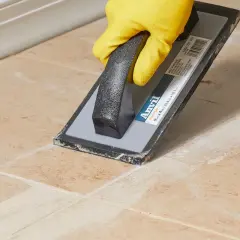 A gloved hand uses a grout float to spread grout, leaving a light grout haze on the tiled surface.