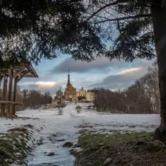 Śnieżny krajobraz z klasztorem na wzgórzu, drzewem i drewnianą wiatą z krzyżem.