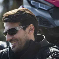 Uomo sorridente con occhiali da moto sportivi, con una moto sullo sfondo.