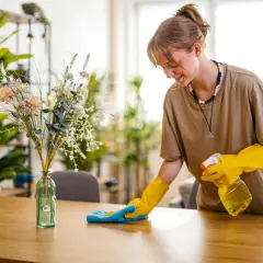 Joven sonríe mientras aprende como limpiar el polvo de los muebles con spray y paño azul.