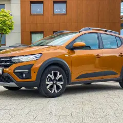Pomarańczowy Dacia Sandero Stepway na tle nowoczesnego budynku. Sprawdź atrakcyjne dacia sandero - ceny.