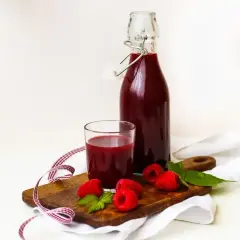 Szklanka i butelka z ciemnoczerwonym sokiem z malin, ozdobione świeżymi malinami i listkami mięty na drewnianej desce.
