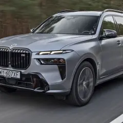 Szary BMW X7 2025 z czarnym grillem i sportowymi akcentami pędzi po drodze wśród drzew.