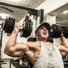 Man med keps och linne lyfter tunga hantlar i ett gym. Han pressar sig till max med varje repetition i sitt cluster set.
