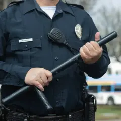 Pracownik ochrony w granatowym mundurze trzyma pałkę. Na piersi widoczna odznaka i mikrofon.