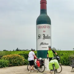 Zwei Radfahrer pausieren vor einer riesigen Weinflasche in Medoc, Frankreich. Weinberge erstrecken sich bis zum Horizont.