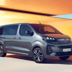 Nowy Peugeot e-Traveller, szary van, zaparkowany przed nowoczesnym budynkiem z żółtymi akcentami.