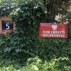 Czerwona tablica z godłem Polski i napisem "DOM EMERYTA WOJSKOWEGO" wśród zieleni. Obok numer domu 5.
