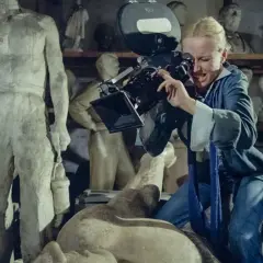 Kobieta z kamerą w pracowni rzeźbiarskiej, jak z filmów Feliksa Falka. W tle antyczne posągi.