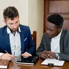 Dwóch mężczyzn w garniturach przy stole, jeden pisze, drugi patrzy na dokumenty.