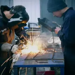Dwóch spawaczy pracuje przy metalowej konstrukcji. Jeden spawa elektrodą, tworząc snop iskier. Drugi obserwuje, trzymając narzędzie.