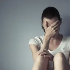 Kobieta w białej koszulce siedzi przy szarej ścianie, zakrywając twarz dłońmi.