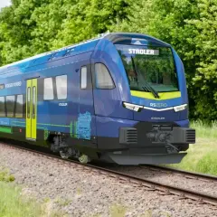 Niebieski pociąg Stadler RSZERO na wodór jedzie po torach wśród zieleni.