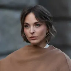 Eine Frau mit Kurzhaarfrisuren und einem leicht molligen Gesicht blickt nachdenklich in die Ferne, eingehüllt in einen braunen Schal.