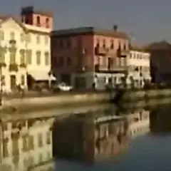 Nadbrzeże z kolorowymi kamienicami odbija się w spokojnej wodzie. W oddali widać fragmenty budynków nad rzeką.