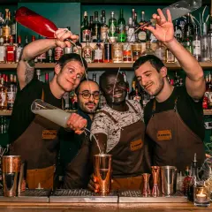 Quatre barmans joyeux préparent des cocktails au bar "Le Renard". L'un verse un liquide d'une bouteille rouge, un autre d'une bouteille transparente.