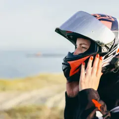 Donna con casco da moto colorato, con mani che aggiustano la visiera per evitare che si appanni. Sfondo sfocato di mare e sabbia.