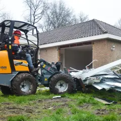 Pomarańczowa koparko-ładowarka z operatorem w kasku i kamizelce usuwa gruz z posesji.