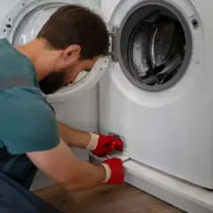 Mężczyzna w czerwonych rękawiczkach otwiera klapkę pralki, by sprawdzić, jak wyczyścić filtr.