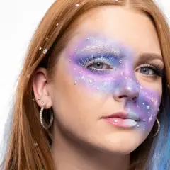En ung kvinna med fantasifull sminkning i lila och blått, inspirerad av 60 tals smink. Glitter och stenar pryder ansiktet.