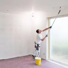 Maler streicht die Decke mit einer Teleskopstange. Ein gelber Farbeimer steht bereit.