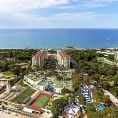 Widok z lotu ptaka na kompleks hotelowy z basenami, zjeżdżalniami i boiskami sportowymi, otoczony zielenią, nad turkusowym morzem. Idealne miejsce na wakacje w Turcji.