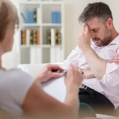 Mężczyzna w jasnej koszuli siedzi z ręką na czole, rozmawiając z terapeutką.