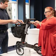 Mann erklärt einer Frau einen Rollator. Eine medizinische Hilfsmittel Liste ist im Hintergrund zu sehen.