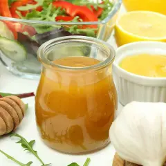 En burk med gyllene salladsdressing, bredvid en skål med sallad, citron, vitlök och honung. Ett perfekt salladsdressing recept!