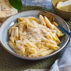 Piatto di penne cremose, condite con una ricca salsa di spaghetti ai 4 formaggi, guarnite con parmigiano grattugiato. Accanto, pezzi di formaggio fresco e stagionato.