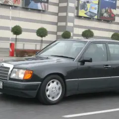 Ciemnoszary Mercedes W124 jedzie ulicą przed budynkiem z banerami.
