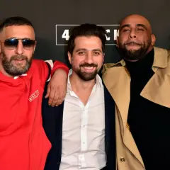 Drei Männer posieren vor einem Banner mit "4 Blocks". Einer trägt eine rote Kapuzenjacke, der andere einen beigen Trenchcoat.