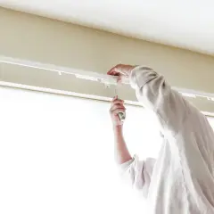 Osoba przykręca śrubokrętem biały karnisz sufitowy nad oknem.
