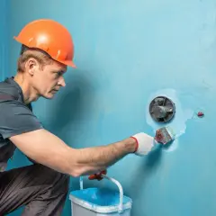 Budowlaniec w kasku maluje pędzlem otwór w niebieskiej ścianie.