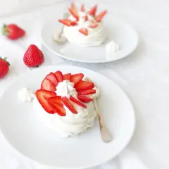 Deux pavlovas à la fraise, légers et aériens, ornés de fruits rouges frais et de crème fouettée, sur des assiettes blanches.