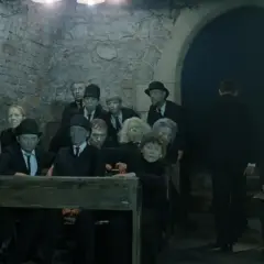 Postacie z "Umarłej klasy" Tadeusza Kantora, w tym uczniowie i nauczyciele, siedzą w ponurej scenerii.