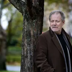 Harald Bovallius lutar sig mot ett träd i en park, klädd i brun rock och halsduk.
