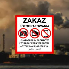 Tablica z napisem "ZAKAZ FOTOGRAFOWANIA" i symbolami przekreślonych aparatów, kamer i telefonów komórkowych. W tle widać przemysłowy krajobraz z kominami fabrycznymi i statkami. To znak informujący, czego nie można fotografować w Polsce.