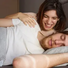 Ein Mann schläft, während eine Frau ihn lachend kitzelt. Vielleicht redet er im Schlaf und sie amüsiert sich.