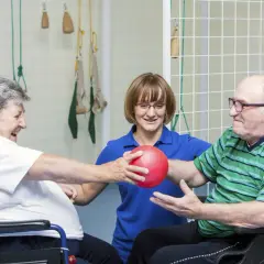 W centrum rehabilitacyjno opiekuńczym dla osób przewlekle chorych somatycznie, pacjenci ćwiczą z fizjoterapeutką, podając sobie czerwoną piłkę.