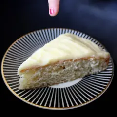 En bit sockerkaka med glasyr på en randig tallrik. En hand med rosa nagellack sträcker sig mot kakan.