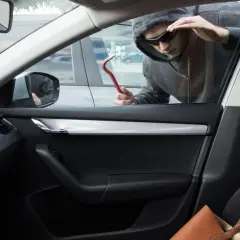 Un homme en capuche et lunettes de soleil tente un vol à la roulotte, outil rouge à la main, regardant à l'intérieur d'une voiture.