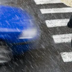 Niebieski samochód przejeżdża obok osoby z parasolem na przejściu dla pieszych podczas deszczu.
