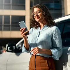 Kobieta w okularach przegląda dane w telefonie, opierając się o samochód. Może to być związane z systemem informacji kredytowej.