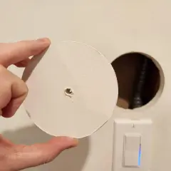 Un disco di cartongesso viene tenuto in mano vicino a un buco nel muro, con un interruttore della luce visibile sotto.