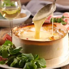 Un Mont d'Or fondant est servi avec une cuillère, accompagné de charcuterie, de pommes de terre et d'une salade verte. Un verre de vin blanc complète ce plat gourmand.