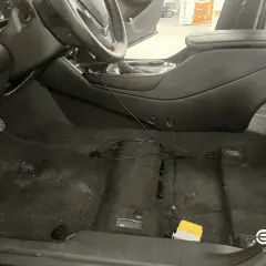 Interior de coche con moqueta oscura y algo de suciedad. Se ve el volante, consola central y pedales. Hay cables y un pequeño objeto amarillo.