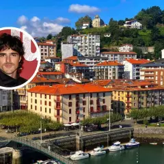 Jacob Elordi, con su aire de galán, posa sobre un pintoresco pueblo vasco con casas de tejados naranjas y un puerto lleno de barcos.