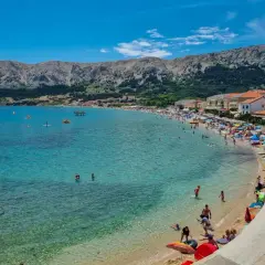 Słoneczna plaża na wyspie Krk, pełna ludzi kąpiących się i opalających. W tle góry i zabudowania. To idealne miejsce na wakacyjne atrakcje.