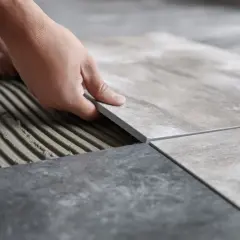 Układanie płytek 60x60. Cennik prac glazurniczych. Dłoń kładzie nową płytkę na kleju.